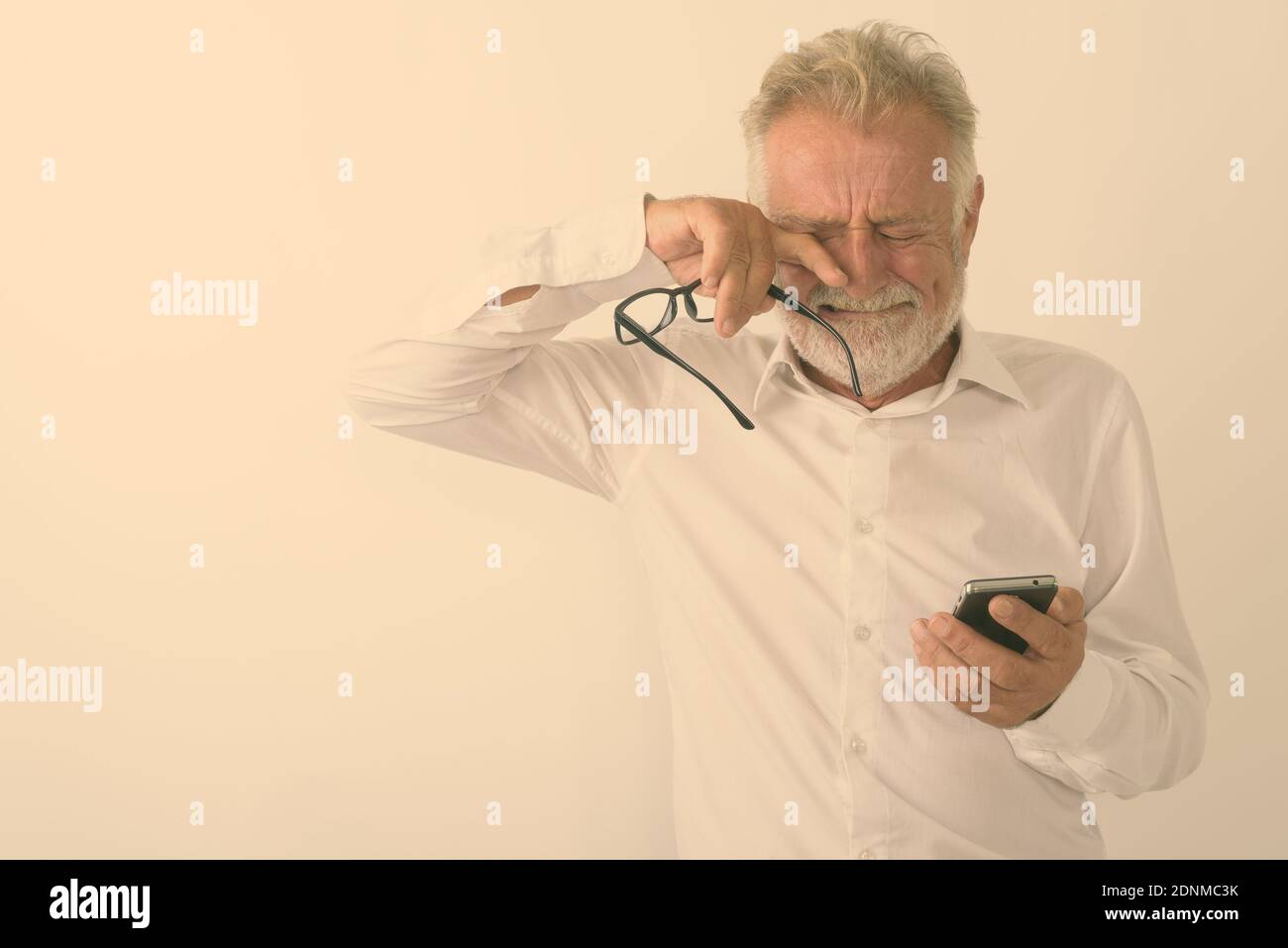 Studio shot of sad senior homme barbu à l'aide de téléphone mobile tout en pleurant et holding eyeglasses against white background Banque D'Images