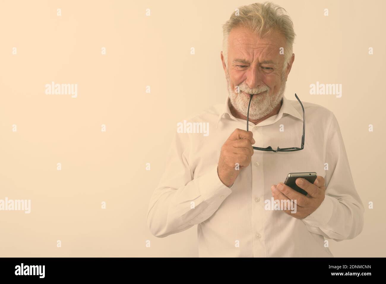 Portrait d'homme barbu senior triste à pleurer pendant l'utilisation de téléphone mobile et de mordre les lunettes contre fond blanc Banque D'Images