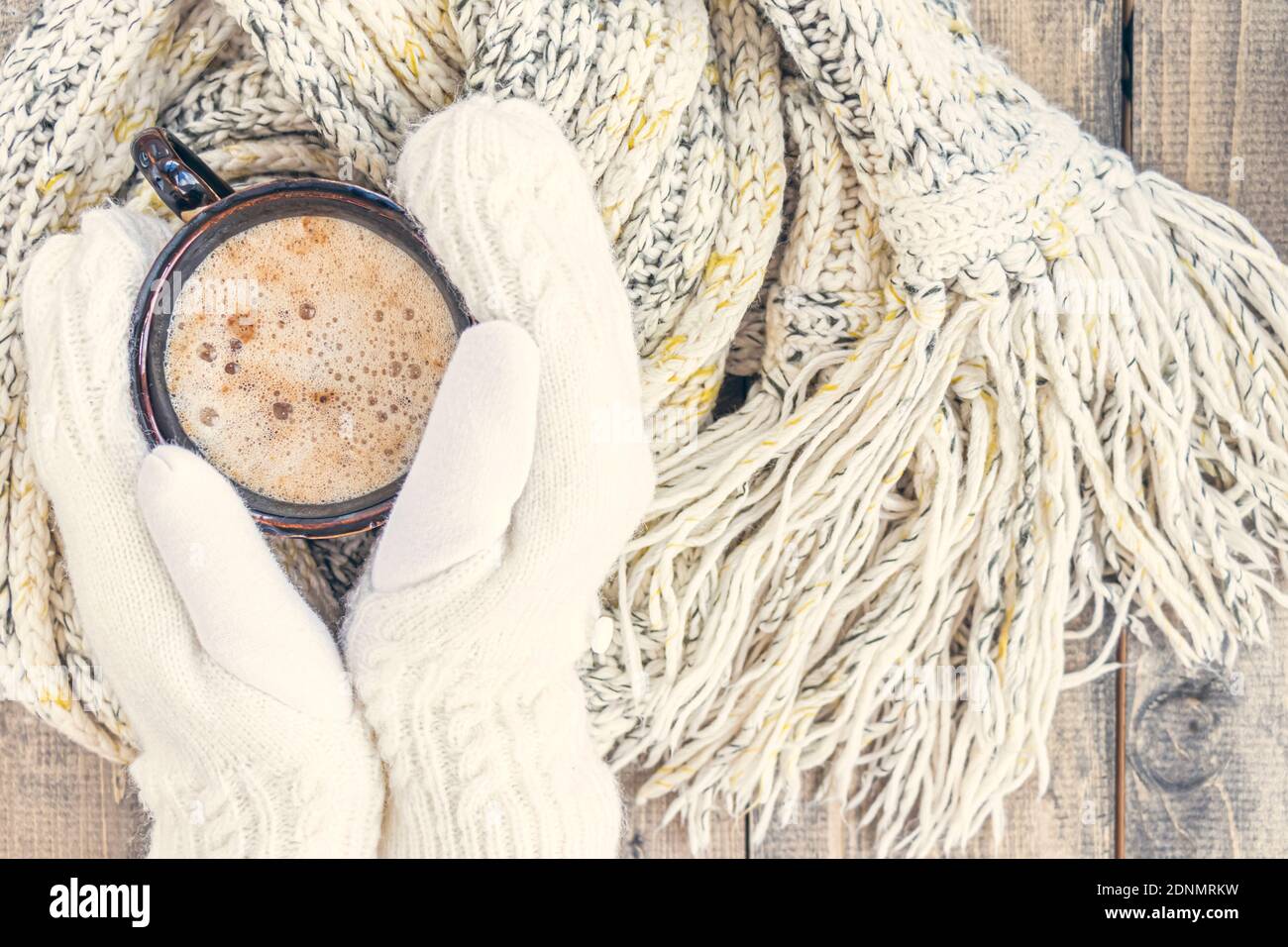 Les mains des femmes en moufles blanches tiennent une tasse de café chaud  avec mousse sur une table en bois avec un foulard en laine à maille chaude.  Arrière-plan de Noël.concept d'hiver,
