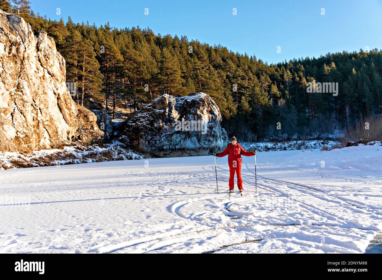 Jeune homme en vêtements d'hiver rouges ski le long de la rivière enneigée gelée près de belles falaises avec forêt de pins. . Activités d'hiver et divertissement, sports, actif Banque D'Images
