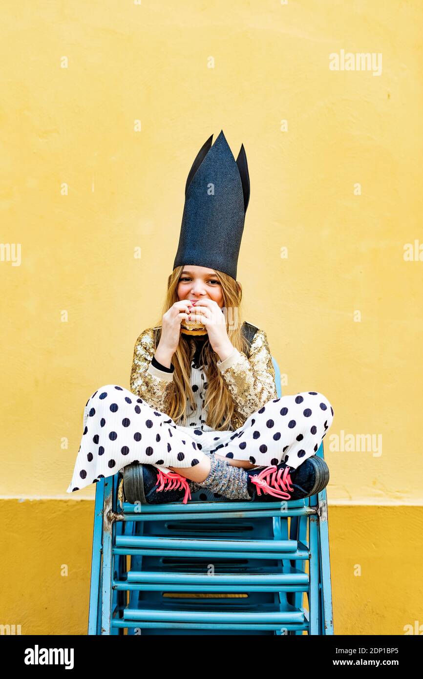 Portrait of Girl wearing black crown assis sur pile de chaises eating Hamburger Banque D'Images