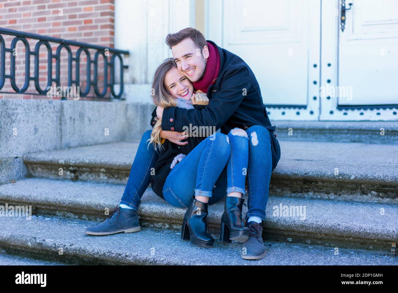 Portrait d'un couple souriant dans des vêtements d'hiver qui s'embrasent assis quelques escaliers sur une scène d'hiver de parc urbain Banque D'Images