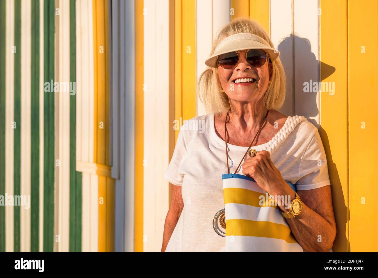 Femme senior à la mode avec lunettes de vue et lunettes de soleil debout devant de mur à rayures colorées Banque D'Images
