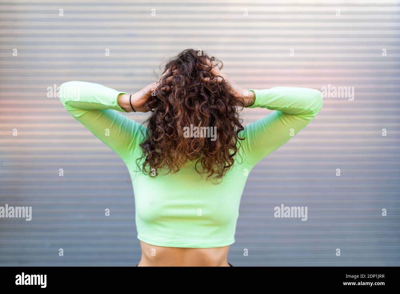 Jeune femme portant un crop top vert avec les mains dans les cheveux poser contre un mur gris Banque D'Images