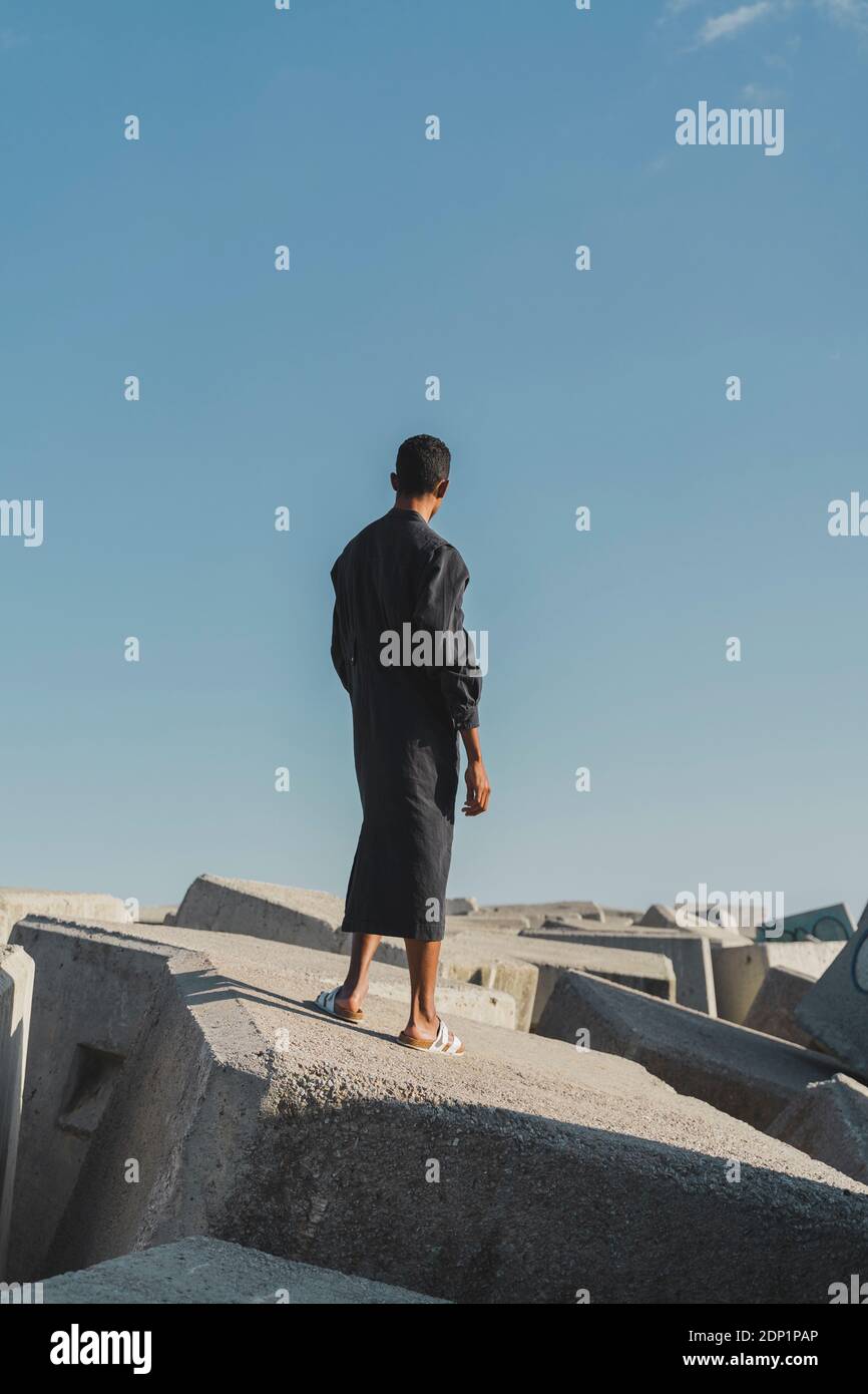 Jeune homme portant un caftan noir debout sur des blocs de béton sous le ciel bleu Banque D'Images