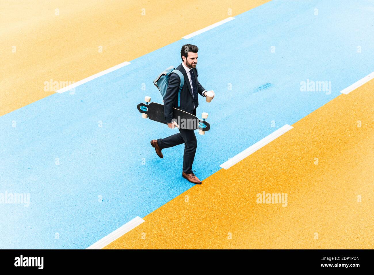 Jeune homme entrepreneur tenant un skateboard et une tasse de café tout en marchant sur la rue jaune et bleue Banque D'Images