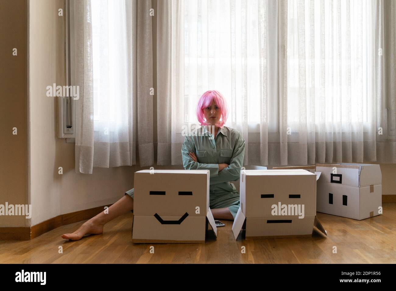 Jeune femme sérieuse portant une perruque rose assise sur le sol avec une boîte en carton pour le visage souriant et mousseur Banque D'Images