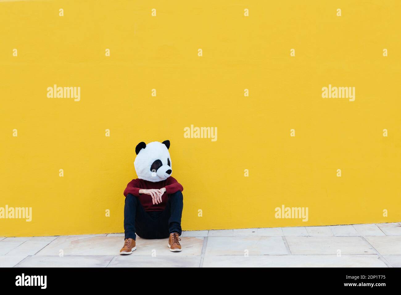 Homme portant un masque de panda tout en étant assis contre le mur jaune Banque D'Images