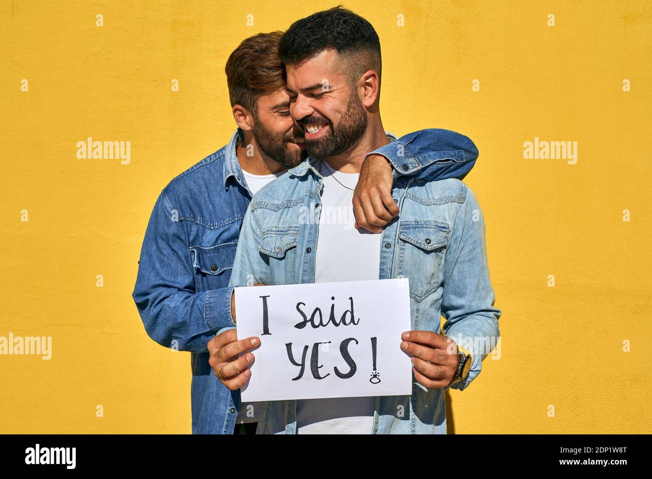Gay homme embrassant jeune homme tenant le papier avec le texte I Dit Oui écrit sur elle contre le mur jaune Banque D'Images