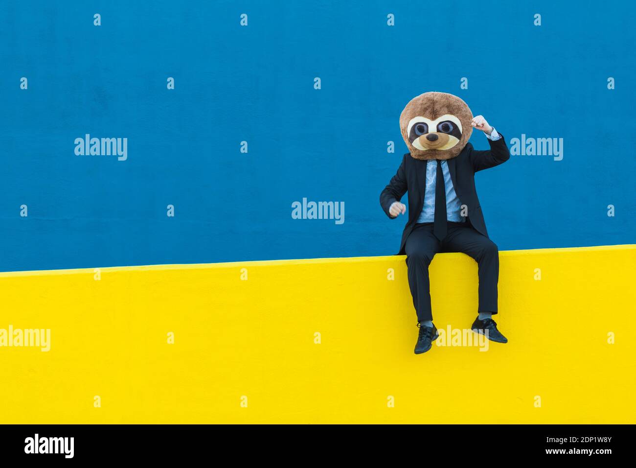Portrait d'homme d'affaires en costume noir avec masque de meerkat assis sur un mur jaune devant un arrière-plan bleu Banque D'Images