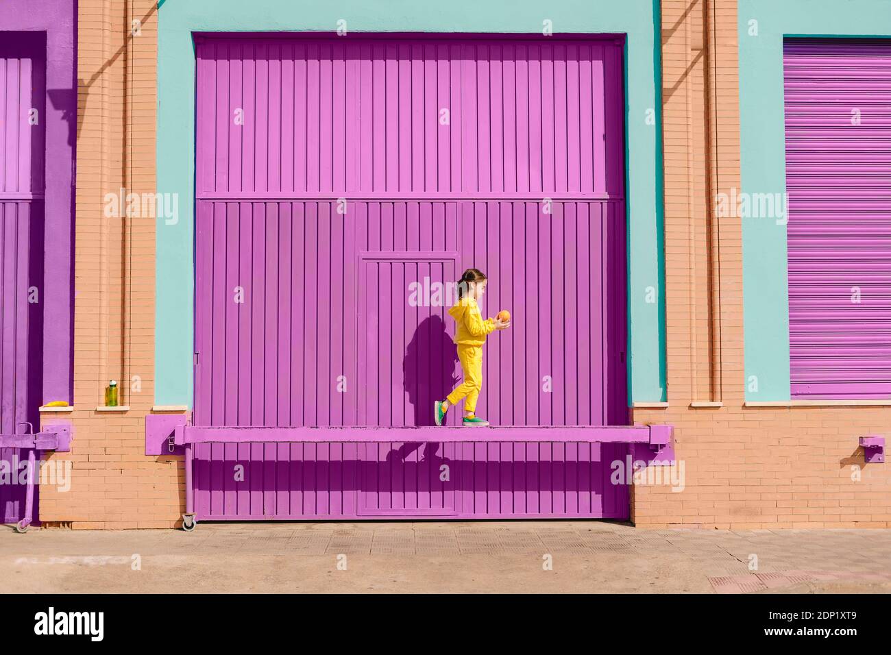 Petite fille vêtue d'un équilibre jaune avec de l'orange sur le bar devant la porte de garage violette Banque D'Images