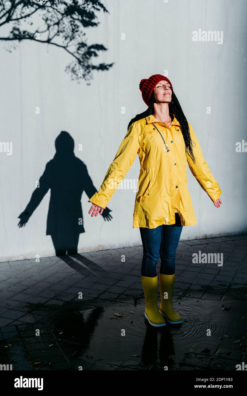 Femme portant un imperméable debout sur une flaque de pluie contre un mur d'ombre Banque D'Images