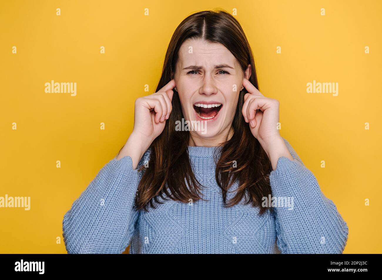 Portrait d'une jeune femme cacaucasienne frustrée et folle couvre les oreilles avec les doigts qui continuent à crier, vêtue d'un chandail bleu Banque D'Images