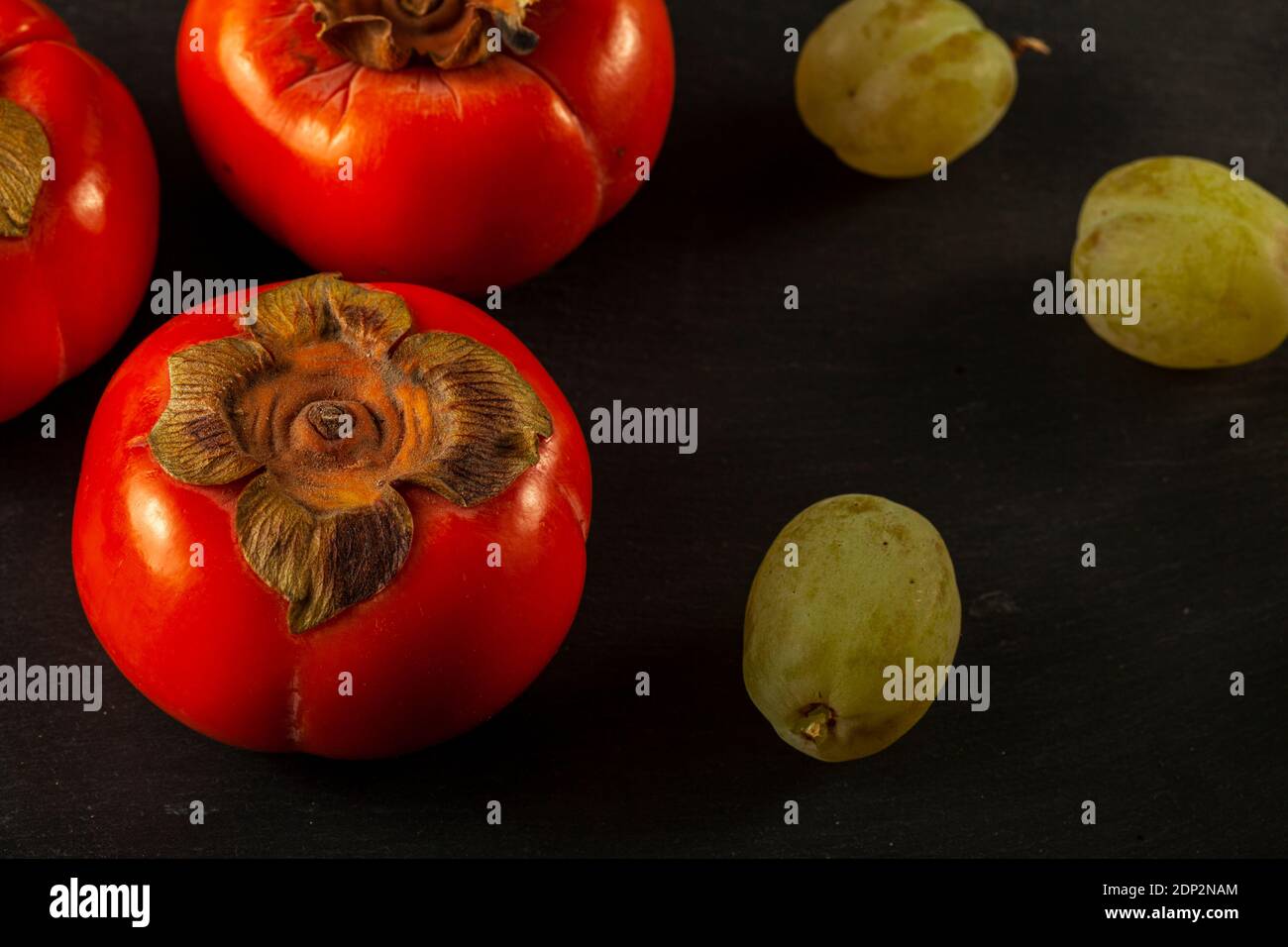 Gros plan de quelques fruits Sharon (persimmons) et quelques raisins verts sur fond noir en bois. Une composition simple de saine alimentation fraîche co Banque D'Images