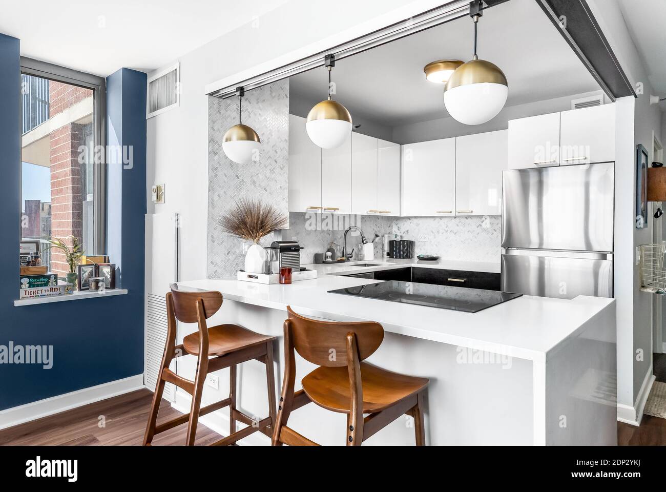 Une cuisine blanche avec des lumières dorées suspendues au plafond, des tabourets de bar assis au comptoir en granit et un mur décoratif bleu. Banque D'Images