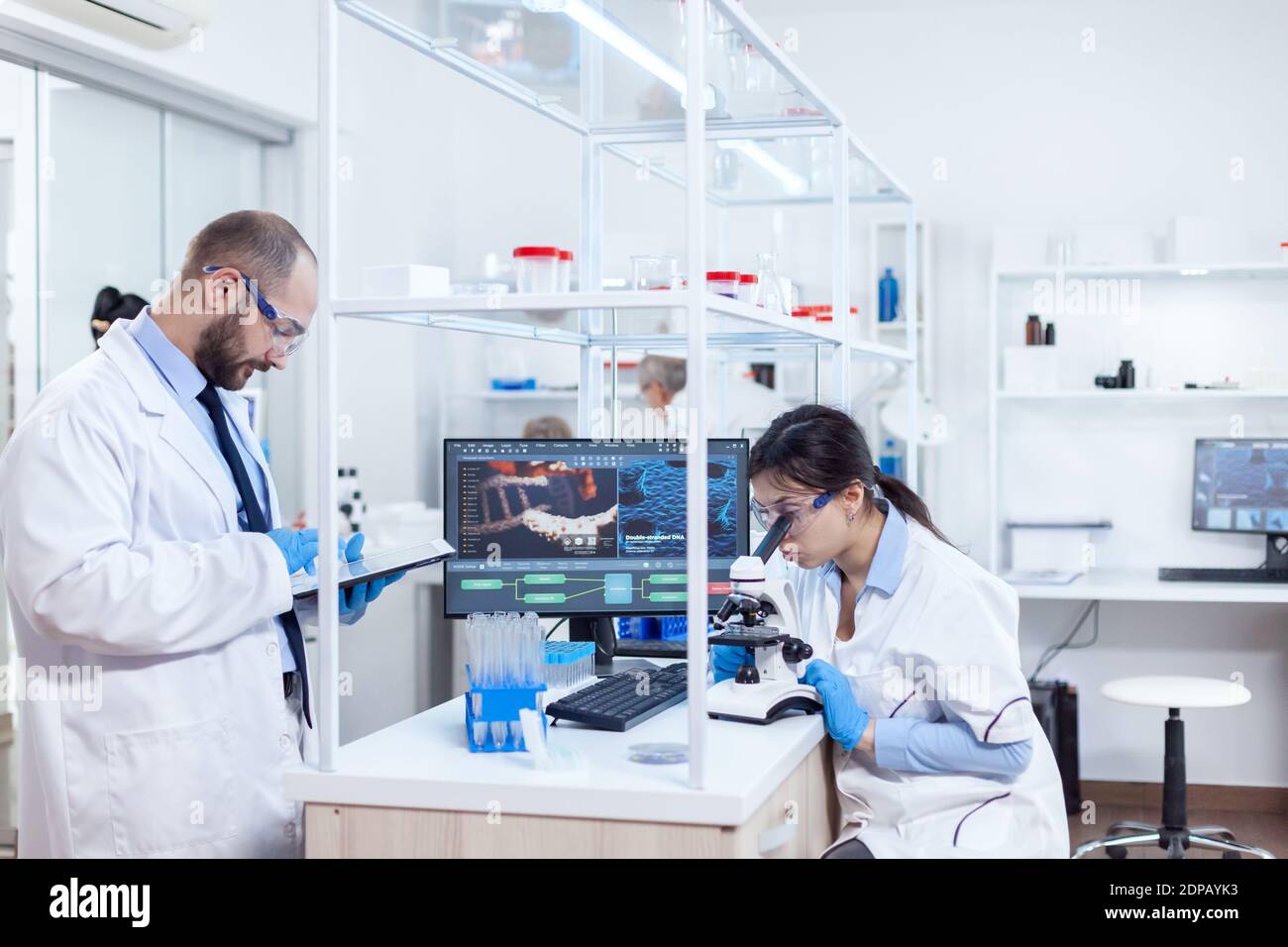 Chimiste portant des lunettes de sécurité et utilisant un Tablet pc en laboratoire. Équipe de chercheurs en génie de la pharmacologie dans un laboratoire stérile pour l'industrie de la santé avec un assistant africain en arrière-plan. Banque D'Images