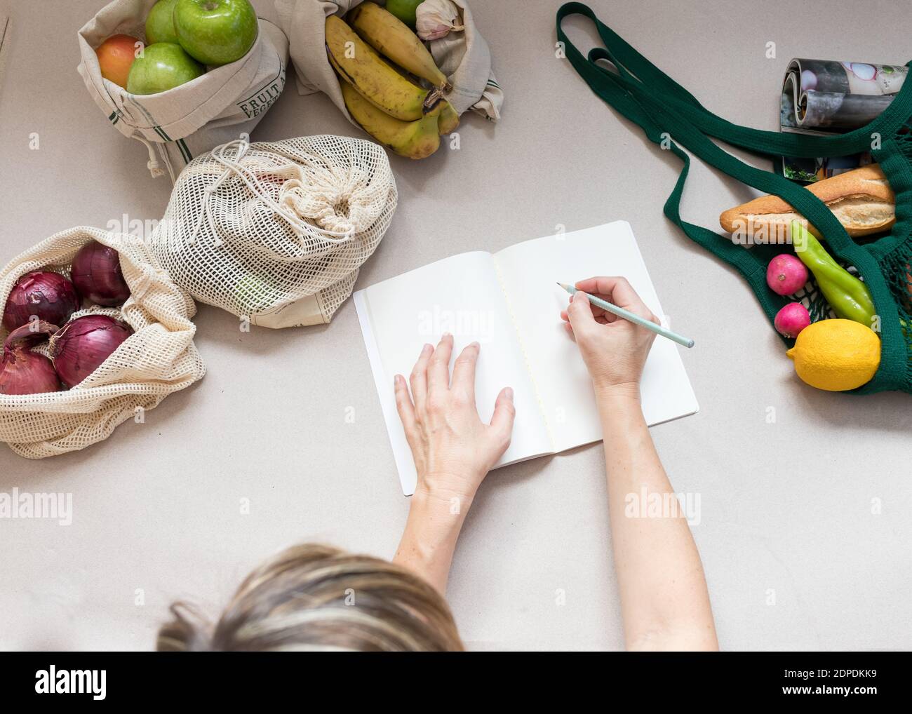 Femme faisant une liste d'achats sur un bloc-notes blanc. Beaucoup de fruits et légumes sains restent à l'intérieur de sacs écologiques durables. Banque D'Images
