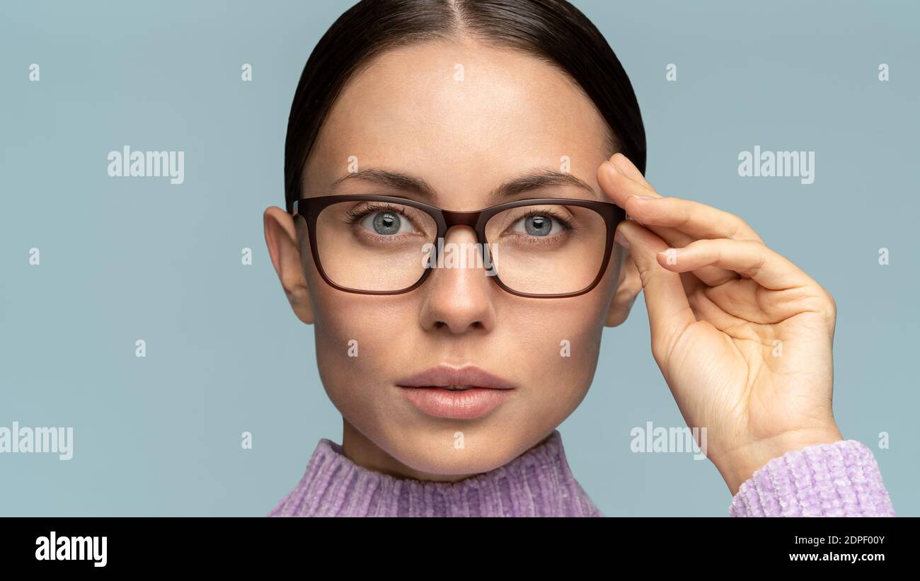 La femme d'affaires sérieuse auto-assurée touche des lunettes, regardant l'appareil photo, isolé sur fond bleu studio. Femme confiante avec des cheveux combed adjus Banque D'Images