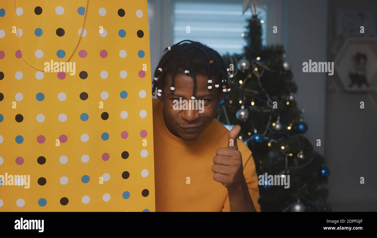 Esprit de Noël. Homme noir afro-américain tenant un grand sac de shopping doré un cadeau devant l'arbre de noël avec le pouce vers le haut. Photo de haute qualité Banque D'Images