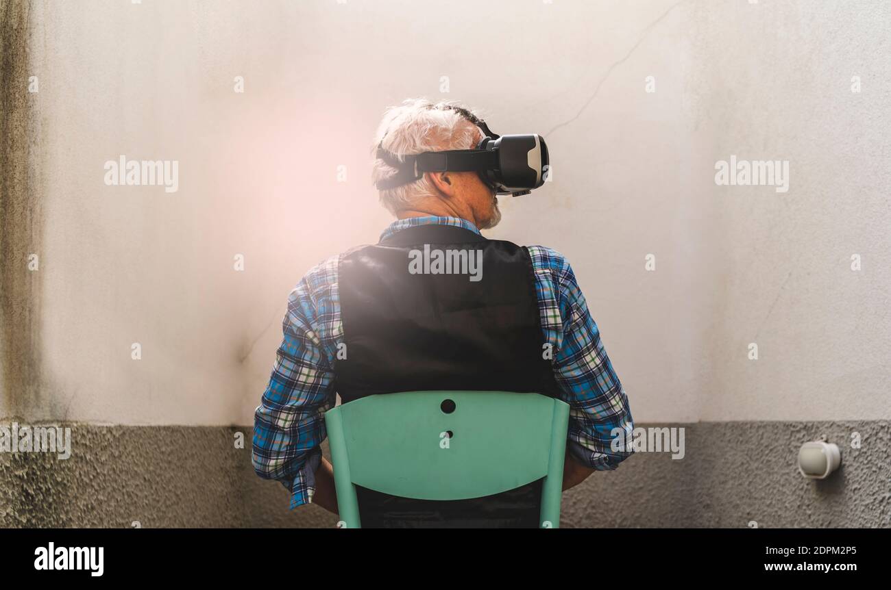 Vue arrière de l'homme senior regardant à travers le simulateur de réalité virtuelle tout en étant assis sur la chaise Banque D'Images