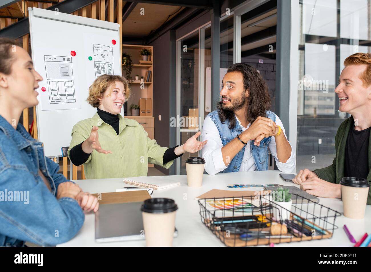 Jeune femme d'affaires joyeuse en vêtements décontractés regardant l'un des hommes collègues avec un sourire éclatant lors de la discussion des idées pour le projet Banque D'Images