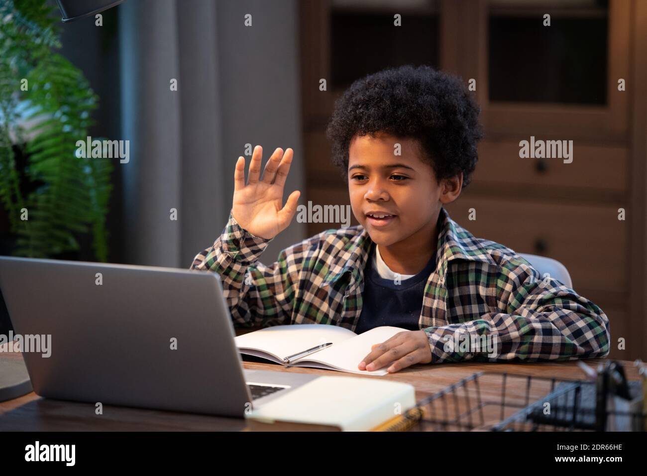 Adorable petit garçon d'école africain en vêtements décontractés, se balayant avec la main tout en étant assis près d'une table devant un ordinateur portable, regardant l'affichage et saluant son professeur Banque D'Images