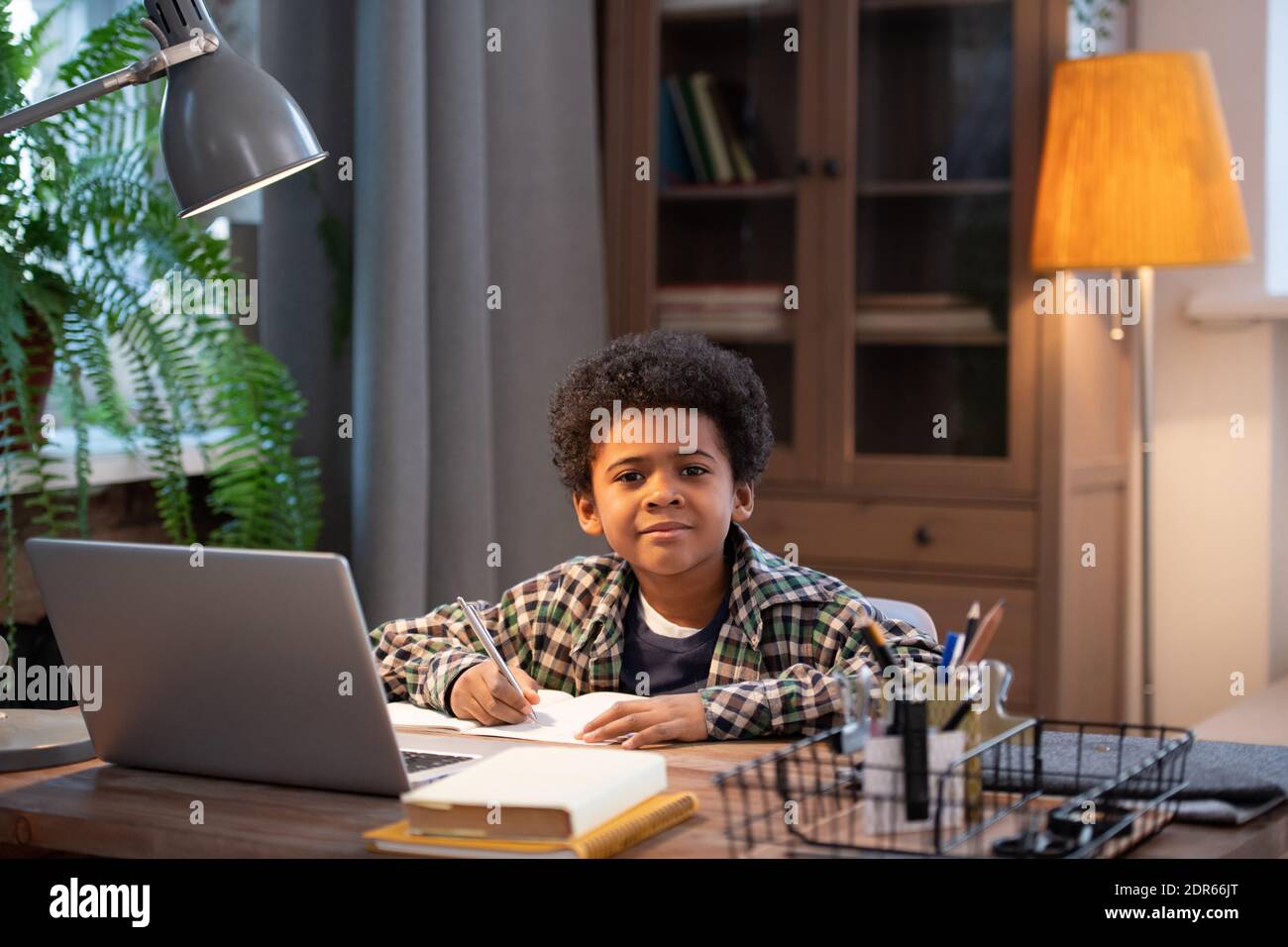 Un écolier africain intelligent et diligent prenant des notes en copybook et vous regardant assis près d'une table devant ordinateur portable dans la salle de séjour Banque D'Images