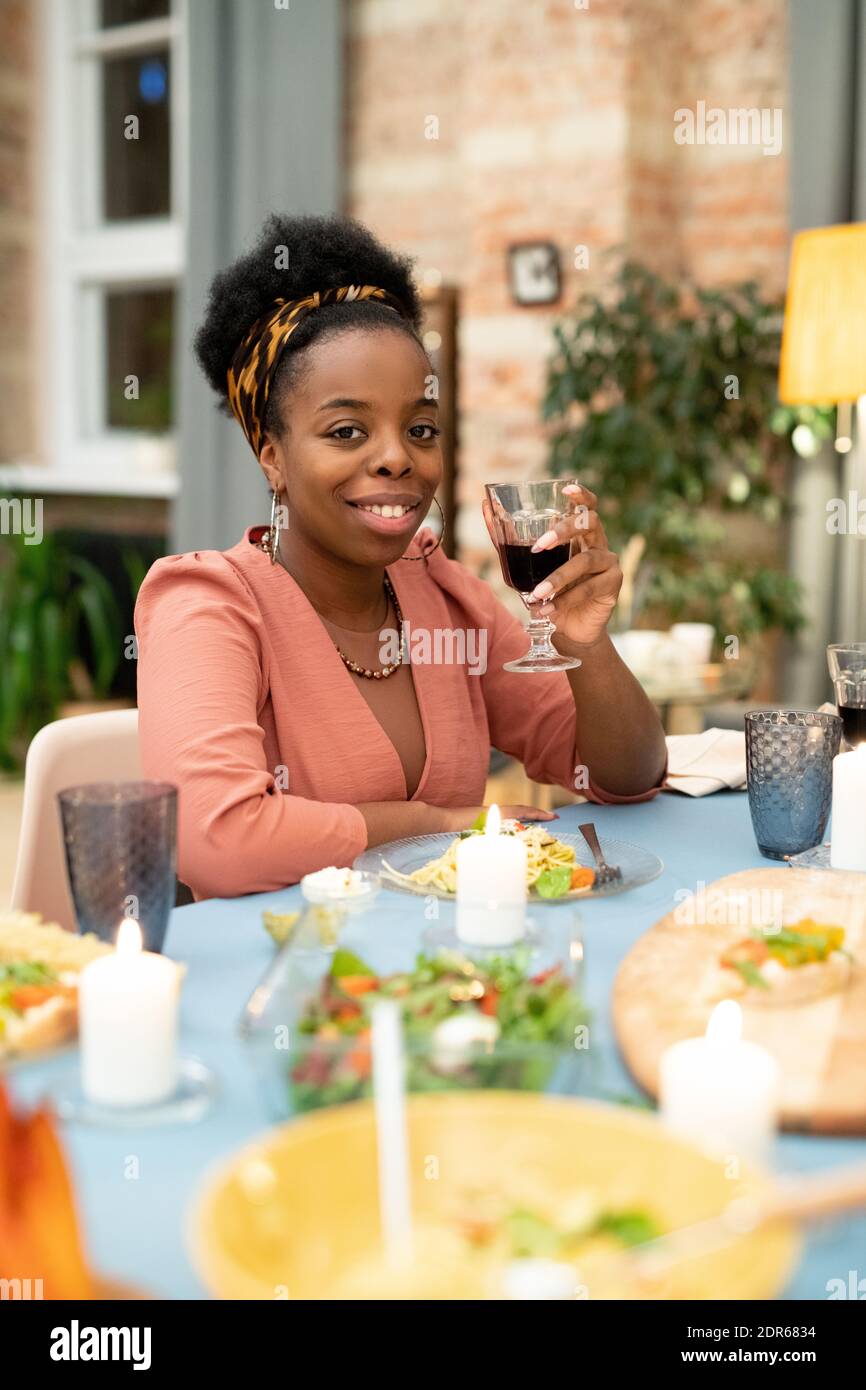 Bonne jeune femme africaine qui applaudisse avec un verre de rouge vin devant l'appareil photo pendant que vous êtes assis près de l'hôtel, servi de fête Table pendant le dîner de Noël Banque D'Images