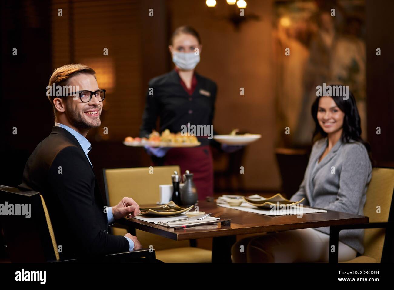 Sympathique gentilhomme et charmante jeune femme souriant et regardant appareil-photo pendant que la serveuse est sous le masque et qu'elle apporte sa commande de nourriture Banque D'Images