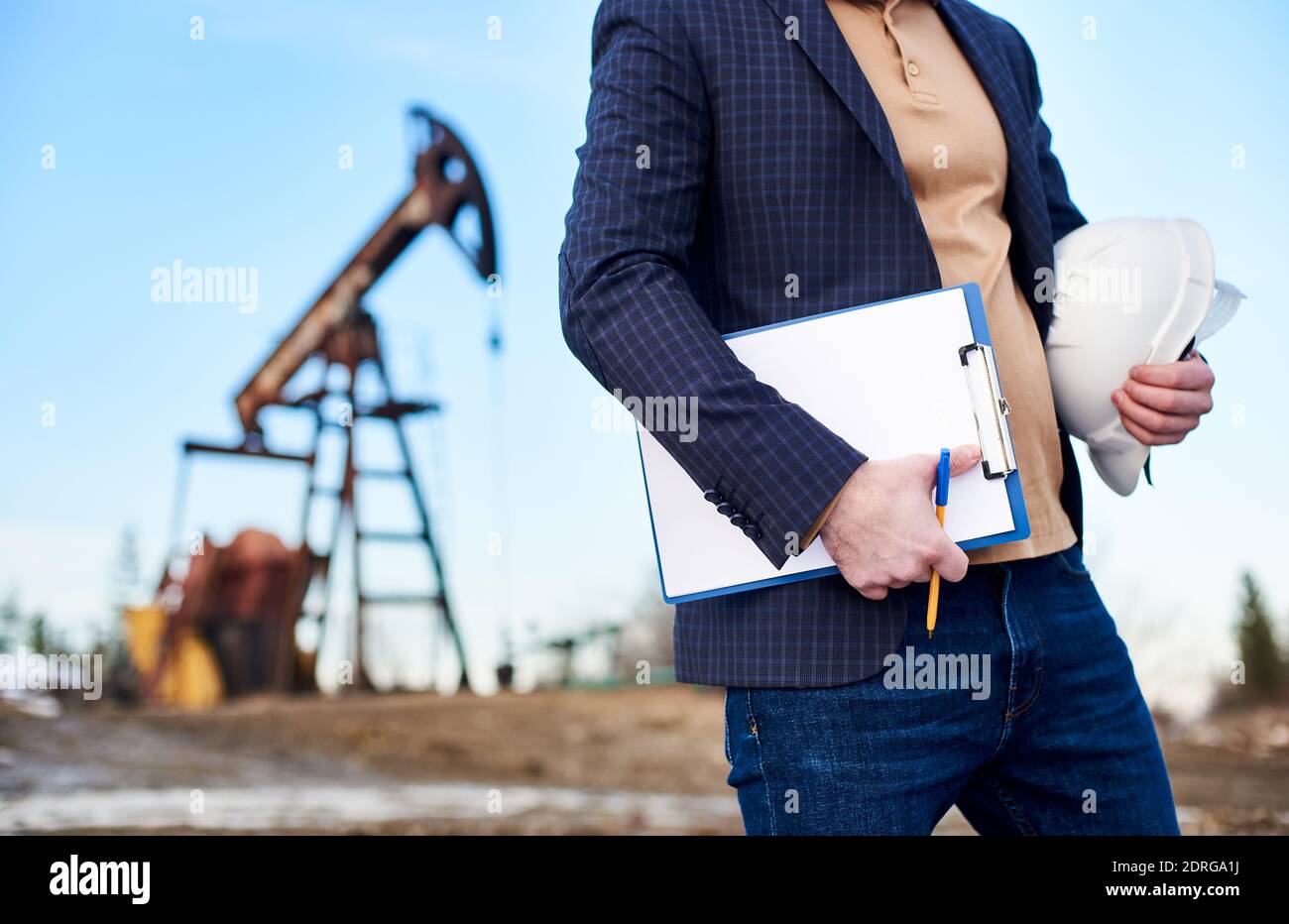 Gros plan d'un homme d'affaires pétrolier tenant un casque de protection, un presse-papiers, un stylo. Pétrolier debout sur le territoire du champ de pétrole avec le cric de pompe sur un fond flou. Concept de l'industrie pétrolière et de la production pétrolière Banque D'Images