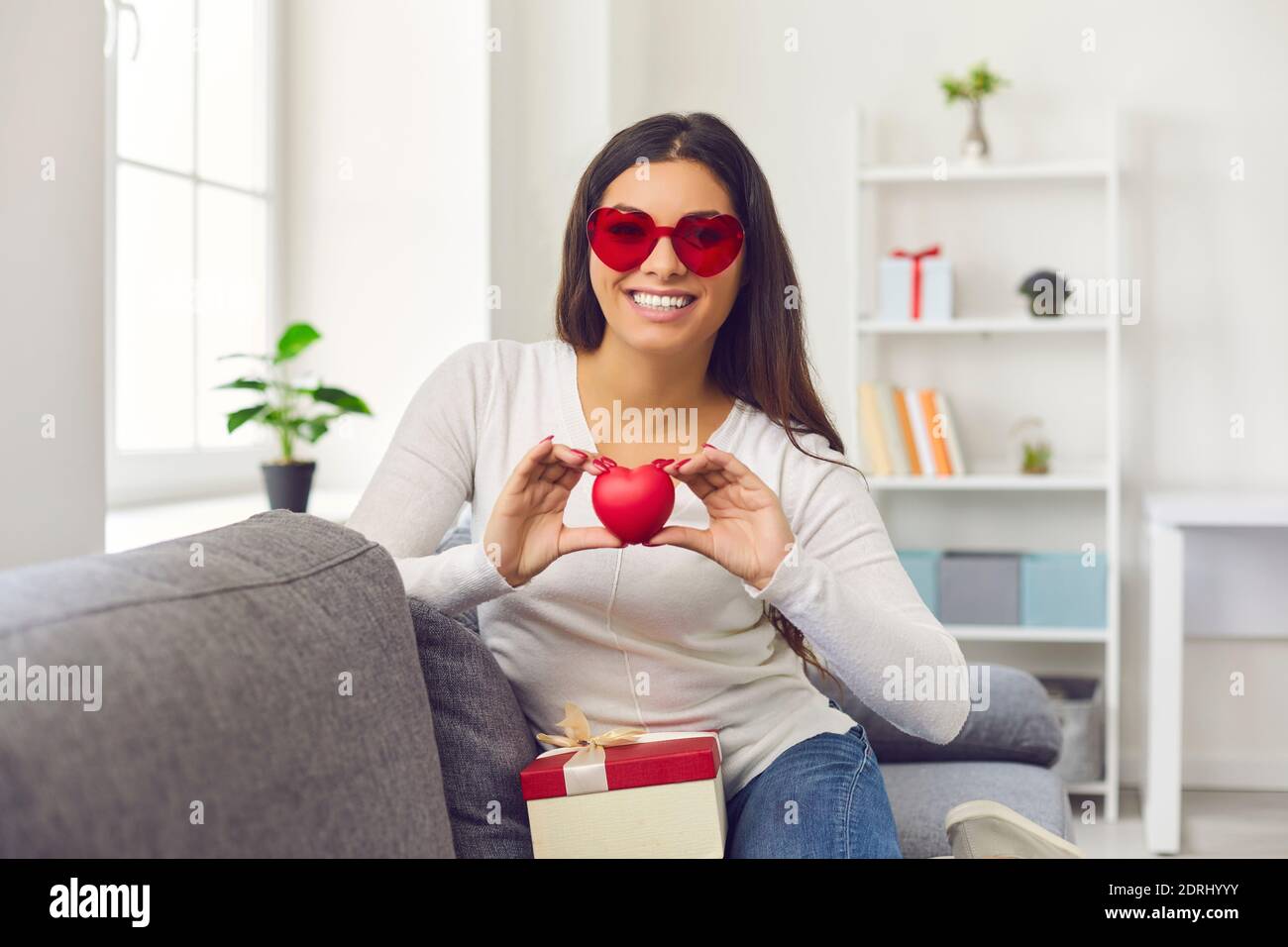 Femme souriante dans des verres en forme de coeur assis sur un canapé avec un cadeau Et tenir le coeur rouge de la Saint-Valentin Banque D'Images