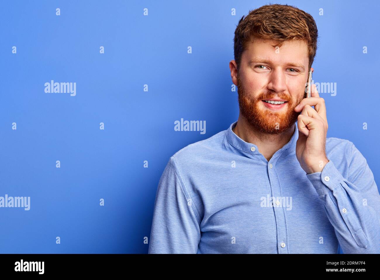 l'homme a une belle conversation avec quelqu'un au téléphone, portant une chemise bleue formelle, ayant des cheveux rouges et la barbe, isolé sur fond bleu Banque D'Images