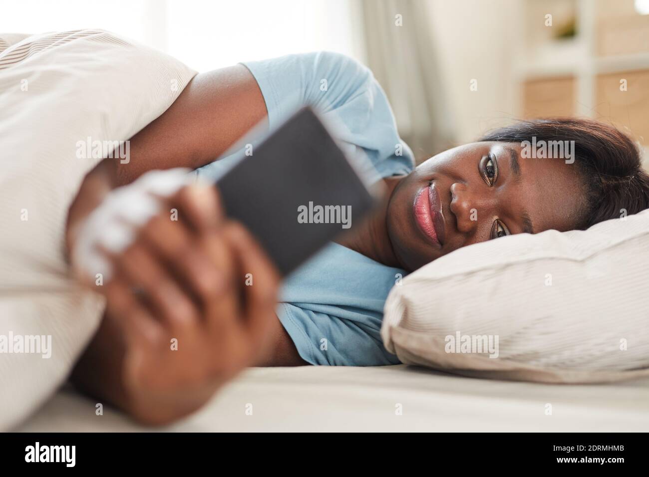 Portrait d'une femme afro-américaine souriante regardant l'écran du smartphone matin, allongé dans un lit confortable Banque D'Images