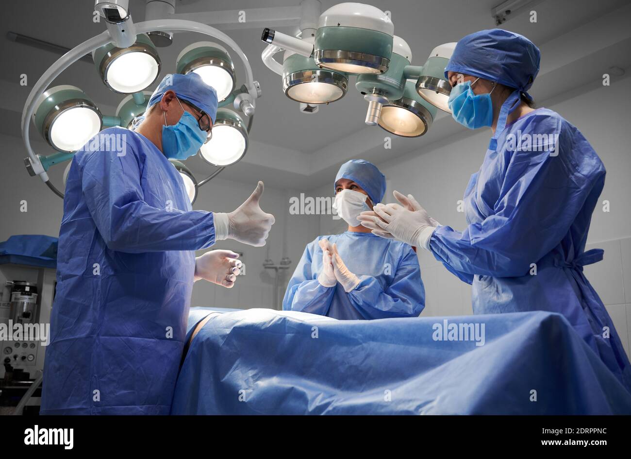 Groupe de médecins en gants stériles et masques protecteurs debout par le patient après une opération chirurgicale réussie. Un chirurgien en plastique mâle fait des pouces vers le haut, tandis que les assistants applaudissent. Banque D'Images