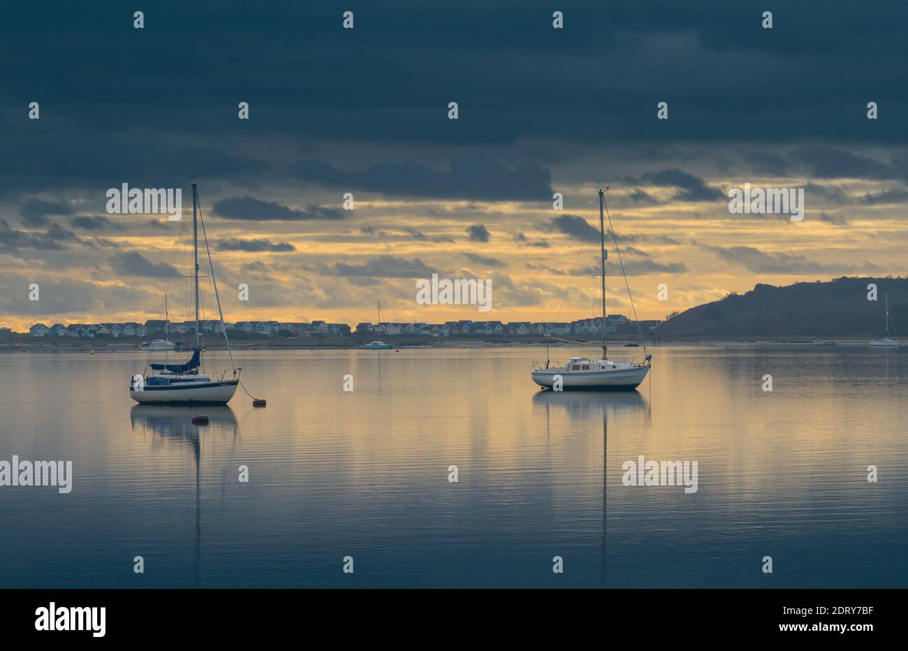 Paire de bateaux à voile avec réflexions à Anchor dans le port de Christchurch avec Mudeford Beach huts en arrière-plan sur UN calme, encore jour au lever du soleil Banque D'Images