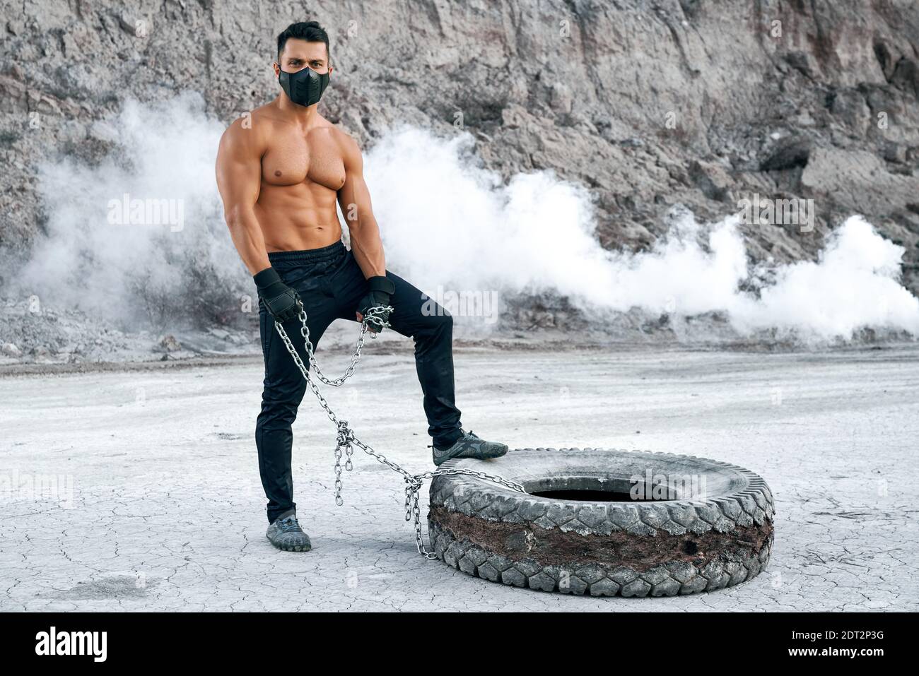 Beau jeune homme avec corps muscle posant a l exterieur tout en se tenant pres de gros pneus. Un homme fort portant un masque noir un pantalon de sport et des gants Photo