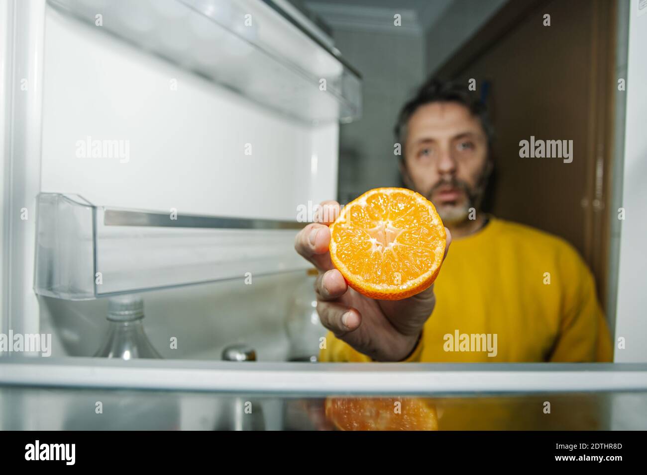 Homme d'âge moyen présentant la moitié de l'orange frais à l'appareil photo en vous tenant près de la porte ouverte du réfrigérateur vide Banque D'Images