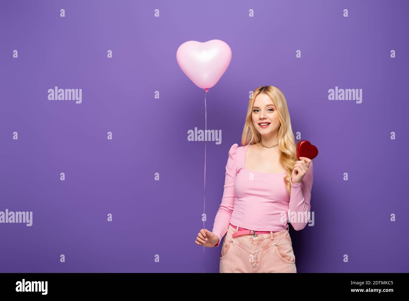 jeune femme blonde souriante tenant un ballon en forme de coeur et un cadeau sur fond violet Banque D'Images