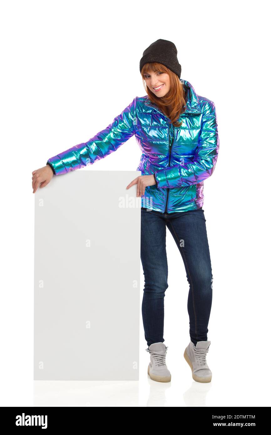 Jeune femme en blouson en duvet, casquette d'hiver, jeans et baskets, pose un écriteau blanc et sourit. Prise de vue en studio sur toute la longueur isolée sur Banque D'Images