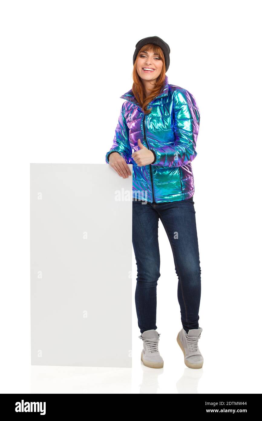 La jeune femme en veste duvet, casquette d'hiver, jeans et baskets est près de l'étiquette blanche, montrant le pouce vers le haut et souriant. Complet le Banque D'Images