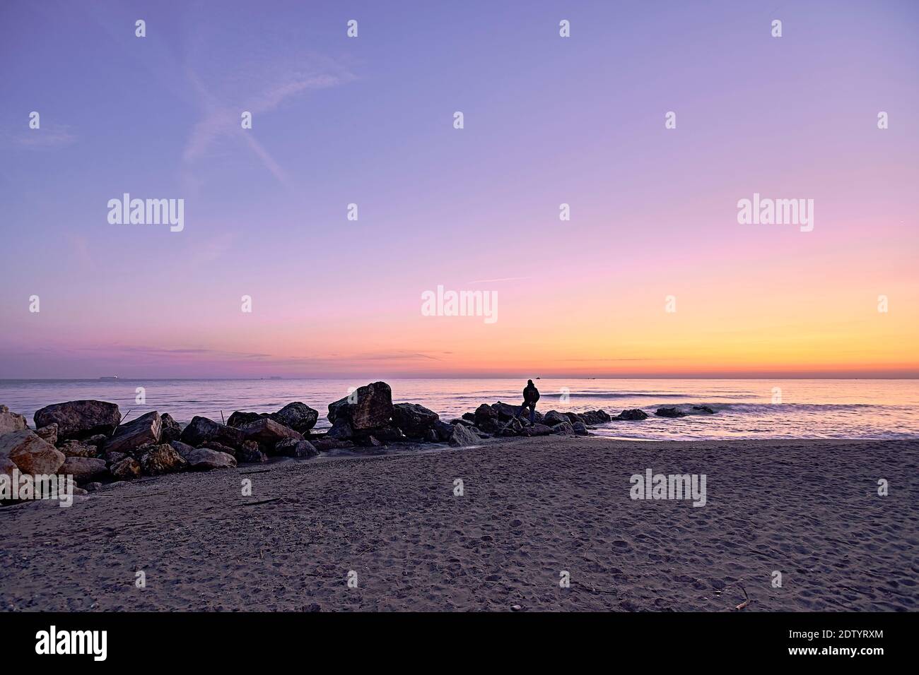 Homme regardant le lever du soleil depuis les rochers sur la plage, solitaire, couleurs orange, sable, mer calme Banque D'Images