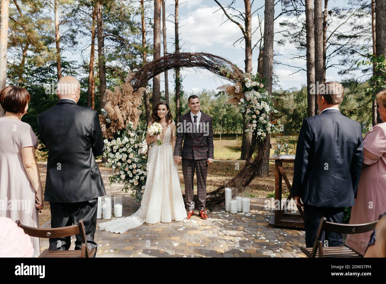 Mariée souriante et marié debout devant l'arche de mariage dans la forêt de pins. Heureux newlyweds à la cérémonie de mariage Banque D'Images