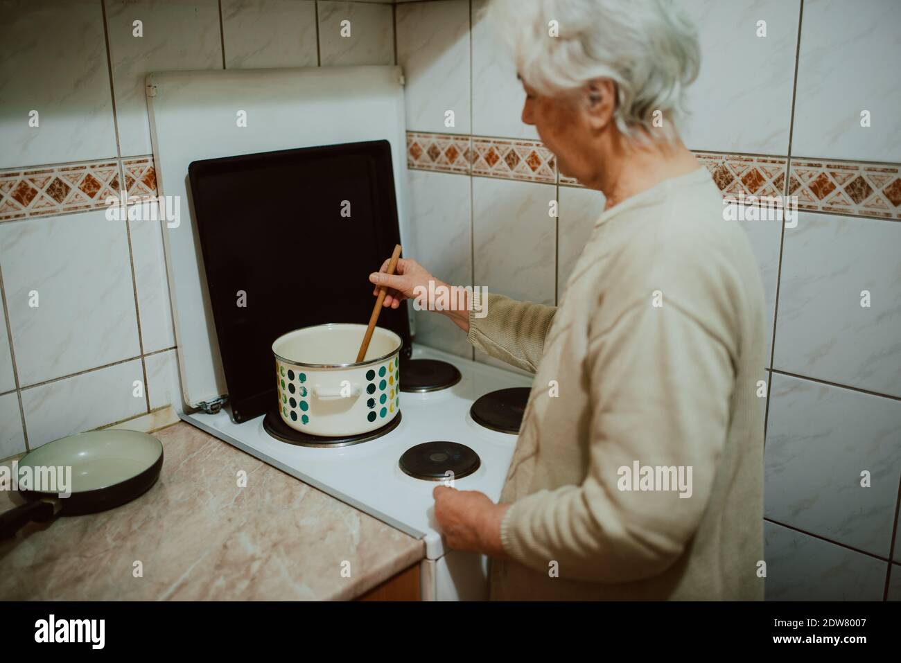Vue de l'arrière d'une femme âgée tout en cuisinant le déjeuner dans la cuisine. Une grand-mère à la retraite aux cheveux gris à côté d'un poêle de cuisine Banque D'Images