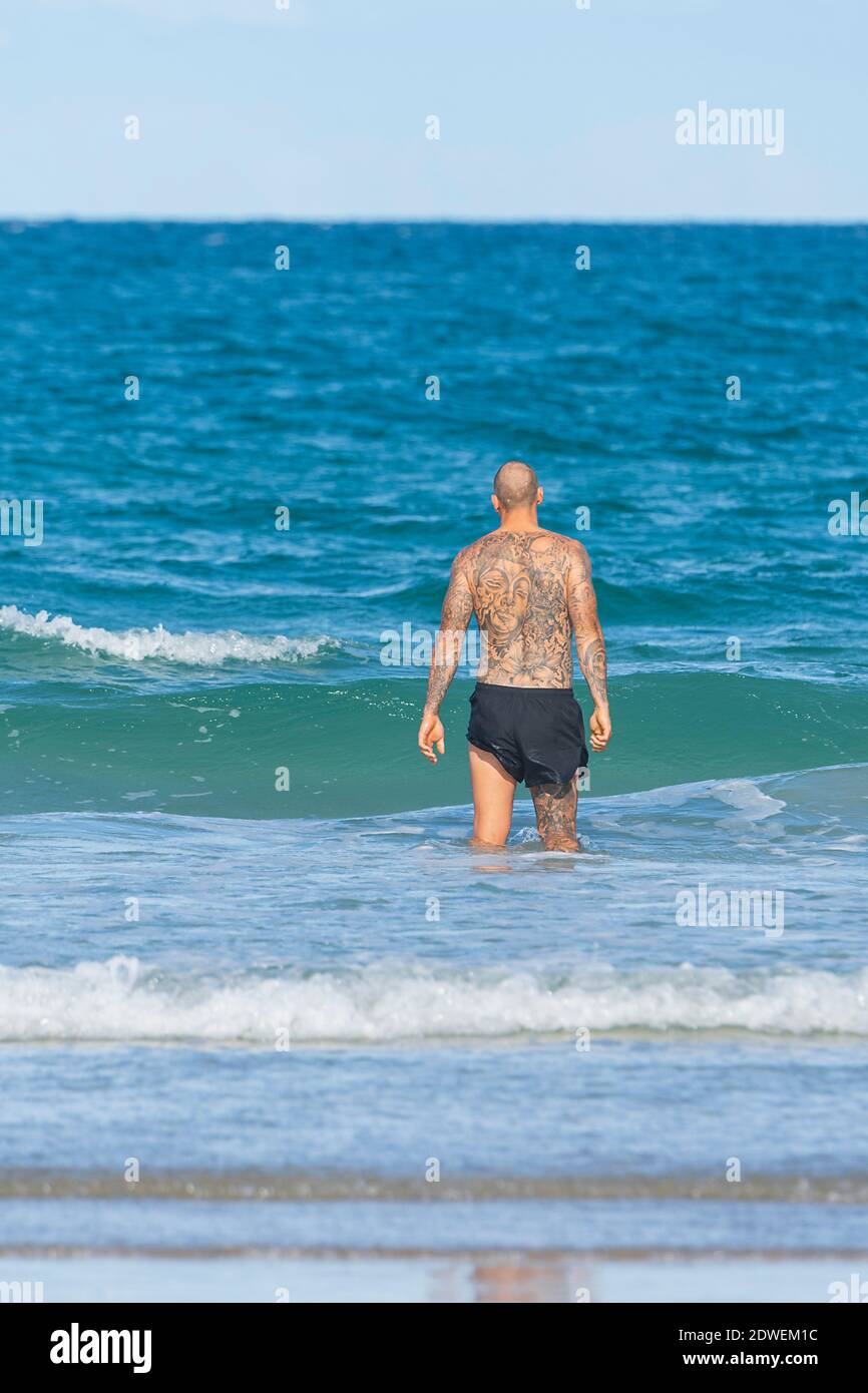 Un homme tatoué se promenant dans la mer sur la plage populaire d'Elliott Heads, Queensland, Queensland, Australie Banque D'Images