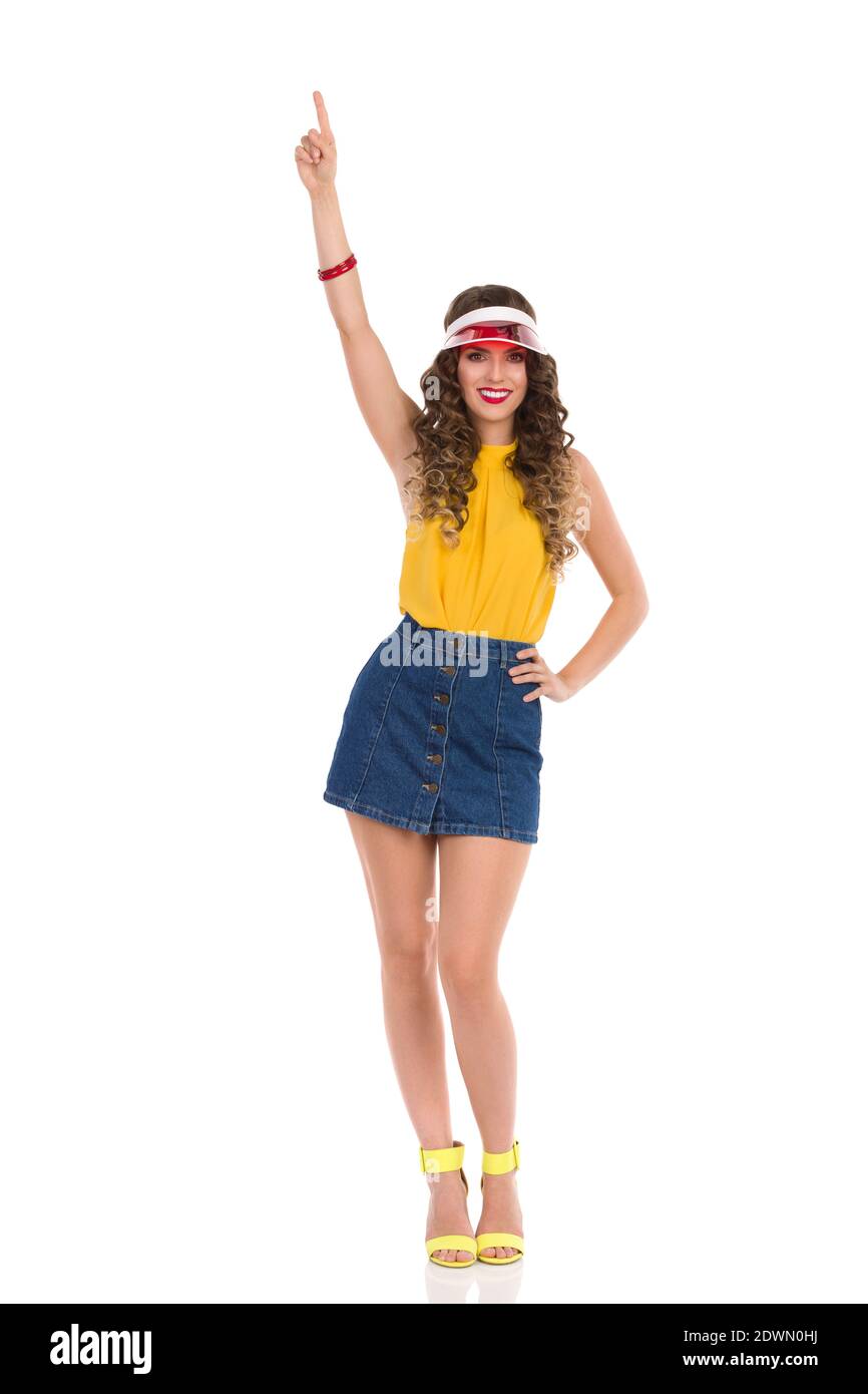 Modèle de mode souriant avec talons hauts sandales, Jean mini jupe, haut jaune et pare-soleil rouge est debout avec le bras relevé et pointant vers le haut. Vue avant. F Banque D'Images