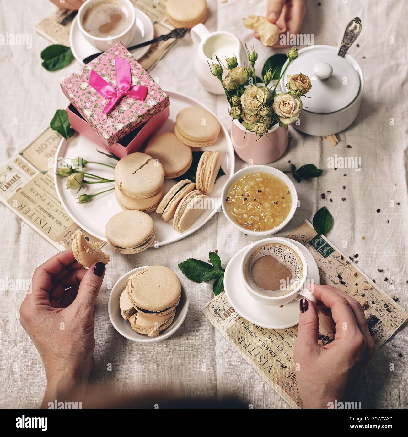 Café au lait, délicieux macarons, pot à lait à la crème, roses et un cadeau sur une nappe en lin et mains féminines gracieuses. Virage affiné. Banque D'Images