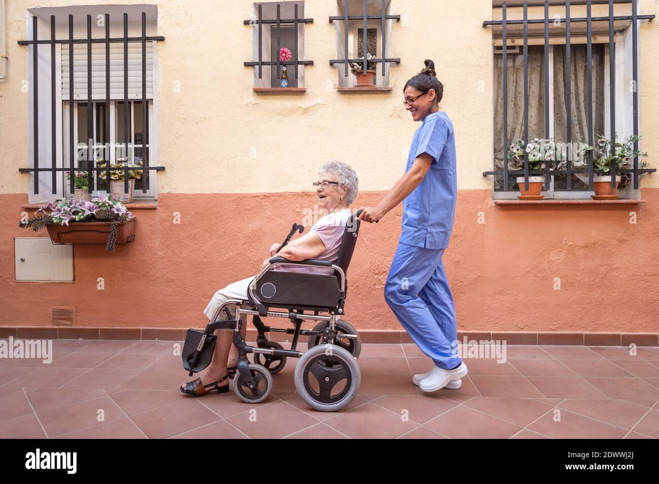 Heureuse femme âgée avec bâton de marche en fauteuil roulant avec son soignant à la maison . Ils parlent et rient ensemble. Banque D'Images