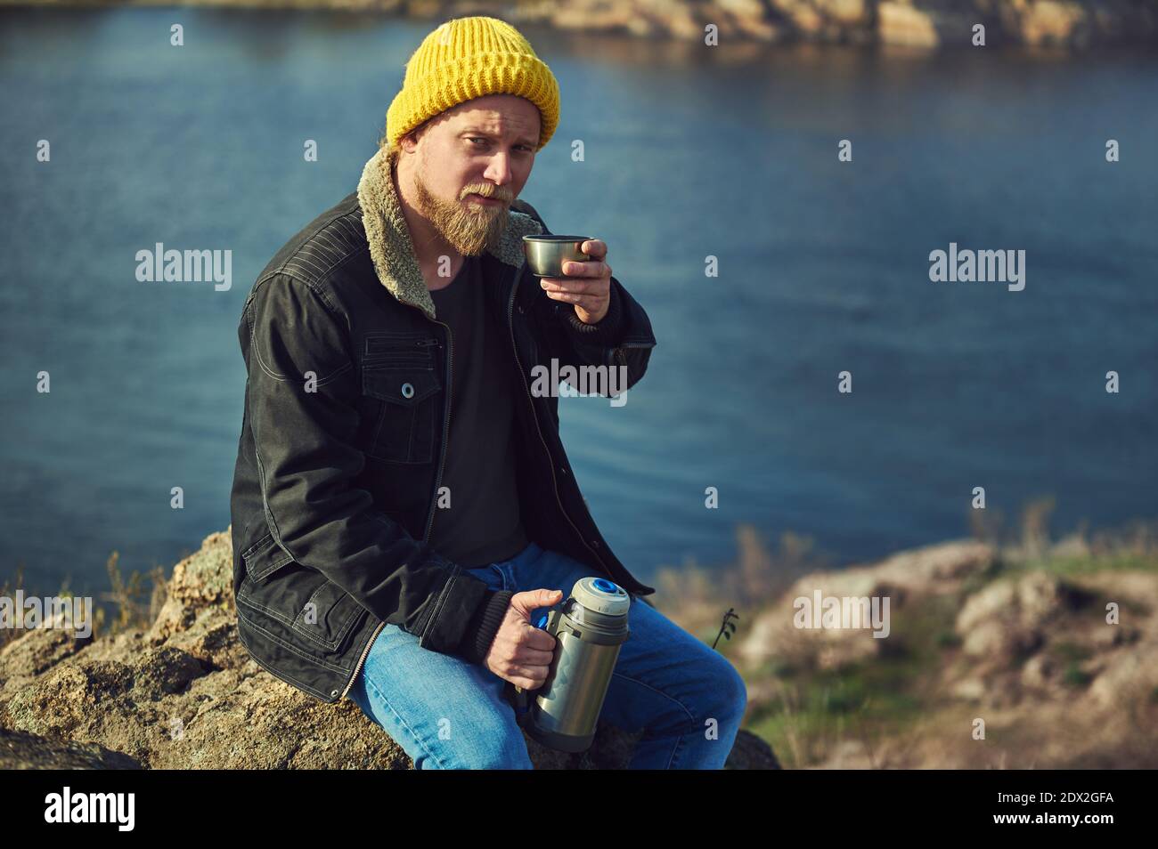 Homme aventureux barbu dans un chapeau jaune tenant un thermos en métal d'une main et d'une tasse de boisson chaude l'autre et faire une pause en randonnée Banque D'Images