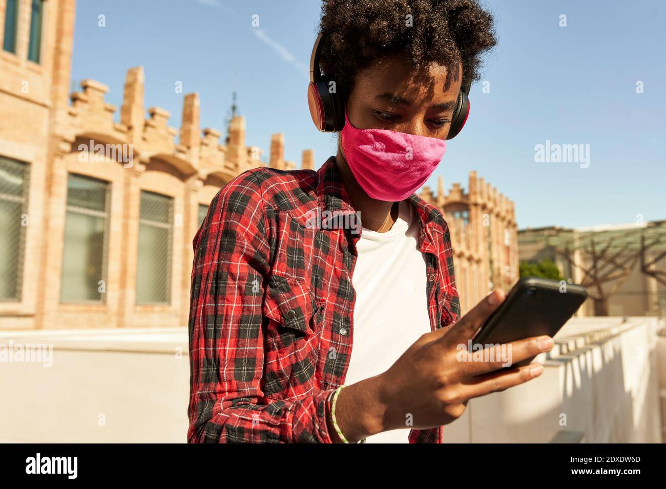 Jeune femme afro portant un masque hygiénique rose tout en utilisant SMART Téléphone pendant COVID-19 Banque D'Images