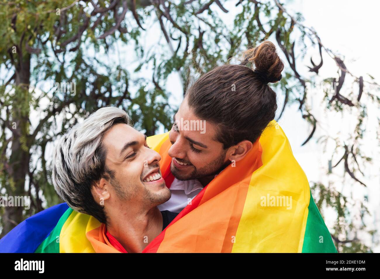 Homme affectueux souriant avec foulard arc-en-ciel, à la recherche d'un partenaire masculin contre l'arbre Banque D'Images